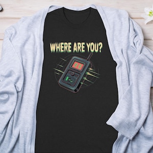 Könnte beinhalten: Schwarzes T-Shirt mit dem Text "WHERE ARE YOU?" und einer Grafik eines Retro-Walkie-Talkies. Das Walkie-Talkie hat ein rotes Display und eine grüne Batterieanzeige. Das Design ist von einem kreisförmigen Muster umgeben.