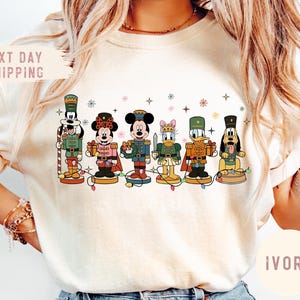 May include: Ivory-colored t-shirt featuring a graphic of Disney characters dressed as nutcrackers. The characters include Mickey Mouse, Minnie Mouse, Donald Duck, and Goofy. The shirt also has the text "Next Day Shipping" and "Ivory".