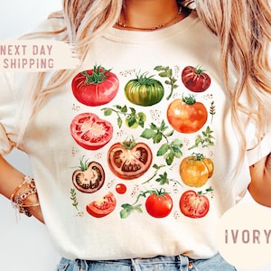 May include: Ivory-colored t-shirt featuring a watercolor illustration of various tomatoes and green foliage. The design includes whole and sliced tomatoes in shades of red, orange, and green. The text "NEXT DAY SHIPPING" is visible on the top left.