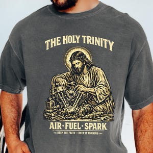 May include: Dark grey t-shirt with a graphic of Jesus working on a motorcycle engine, with the text "THE HOLY TRINITY" above and "AIR FUEL SPARK" below. The shirt also has the text "KEEP THE FAITH - KEEP IT RUNNING".