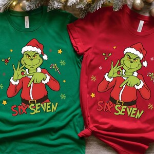 K&ouml;nnte beinhalten: Zwei kurz&auml;rmelige T-Shirts, eines gr&uuml;n und eines rot, mit einer Cartoon-Figur des Grinch mit Weihnachtsm&uuml;tze und rotem Anzug. Die Shirts zeigen den Text "SIX SEVEN" und weihnachtliche Grafiken.