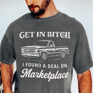 May include: Dark gray t-shirt with white text and a truck graphic. The text reads "GET IN BITCH" above the truck illustration and "I FOUND A DEAL ON Marketplace" below. The shirt is a casual style.