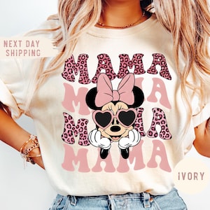 May include: Ivory-colored t-shirt featuring the word "MAMA" in a pink leopard print design, repeated three times. A Minnie Mouse graphic with heart-shaped sunglasses and a pink bow is centered on the shirt. The shirt has short sleeves.