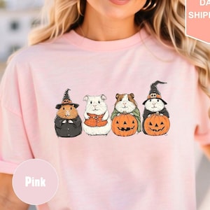 Pode incluir: Camiseta rosa com um gr&aacute;fico de quatro porquinhos-da-&iacute;ndia vestidos com fantasias de Halloween. Os porquinhos-da-&iacute;ndia est&atilde;o usando chap&eacute;us de bruxa, uma echarpe e segurando ab&oacute;boras.