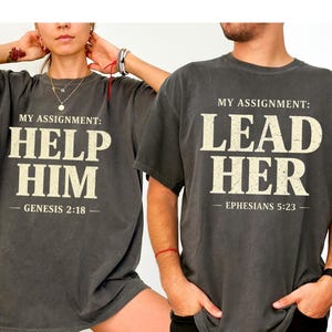 May include: Two gray t-shirts with the text "MY ASSIGNMENT: HELP HIM - GENESIS 2:18" and "MY ASSIGNMENT: LEAD HER - EPHESIANS 5:23" in a bold, vintage-style font. The shirts are worn by two people, and the overall aesthetic is casual and comfortable.