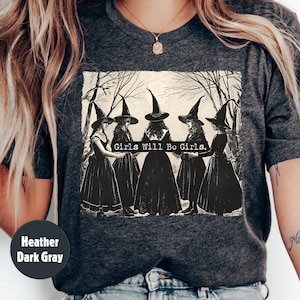 May include: Heather dark gray t-shirt featuring a sepia-toned graphic of five witches in black dresses and hats, standing in a forest. The text "Girls Will Be Girls" is printed above the witches.