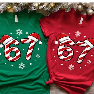 May include: Two short-sleeved Christmas-themed t-shirts, one green and one red. Each shirt features the number "67" designed as a candy cane with a Santa hat and snowflakes. The shirts are displayed on a wooden surface.