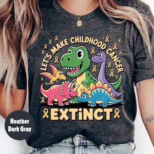 May include: Dark grey heather t-shirt featuring a dinosaur graphic with the text "Let's Make Childhood Cancer Extinct" and yellow ribbons. The shirt has a crew neck design.
