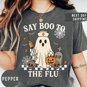 May include: Dark gray t-shirt with a Halloween-themed graphic. The design features a ghost wearing a nurse's hat and holding a syringe, with the text "SAY BOO TO THE FLU". Additional elements include bats, spiderwebs, a pumpkin, and skulls.
