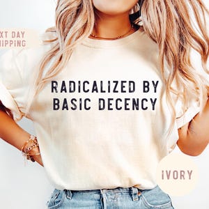 May include: A white t-shirt with the text "Radicalized by Basic Decency" in black lettering. The shirt is being modeled by a person wearing blue jeans. The text "Ivory" is written in a pink circle on the bottom right corner of the image.