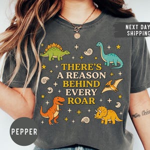 May include: Dark gray t-shirt featuring a colorful dinosaur graphic with the text "THERE'S A REASON BEHIND EVERY ROAR". The design includes various dinosaurs, stars, and shell illustrations. The shirt is a relaxed fit.