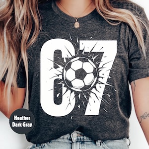 May include: Heather dark gray t-shirt with a large white graphic of the number 67 and a soccer ball. The soccer ball is in the center of the number 67 and has a burst design around it.