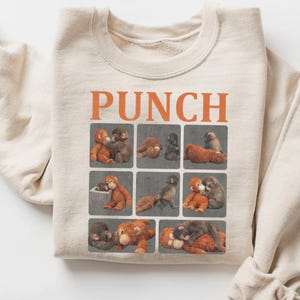 May include: Cream-colored sweatshirt with the word "PUNCH" in orange block letters. The sweatshirt features a grid of nine squares, each with a different illustration of two plush orangutans hugging. The sweatshirt has long sleeves.
