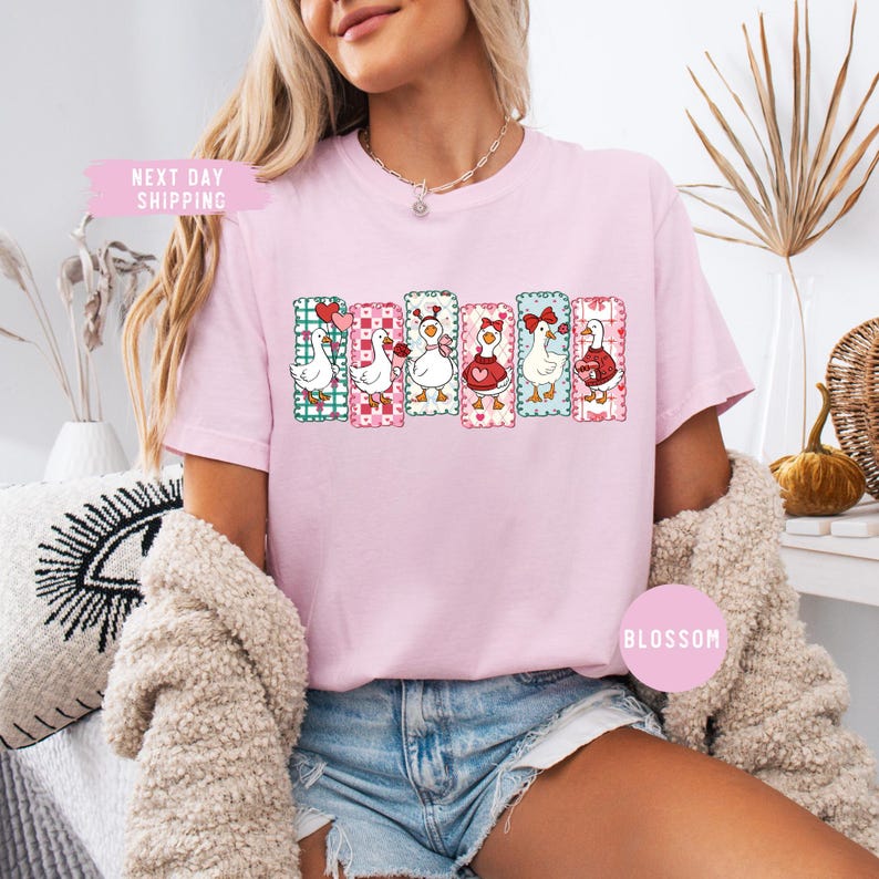 Puede incluir: Camiseta rosa claro con una fila de gansos de dibujos animados, cada uno con dise&ntilde;os de coraz&oacute;n y del D&iacute;a de San Valent&iacute;n. La camiseta tiene la palabra "Blossom" impresa.