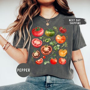 May include: Dark gray t-shirt featuring a watercolor design of various tomatoes in shades of red, green, and orange, with green foliage. The word "PEPPER" is printed on a circle at the bottom left. The words "NEXT DAY SHIPPING" are printed on a black rectangle.