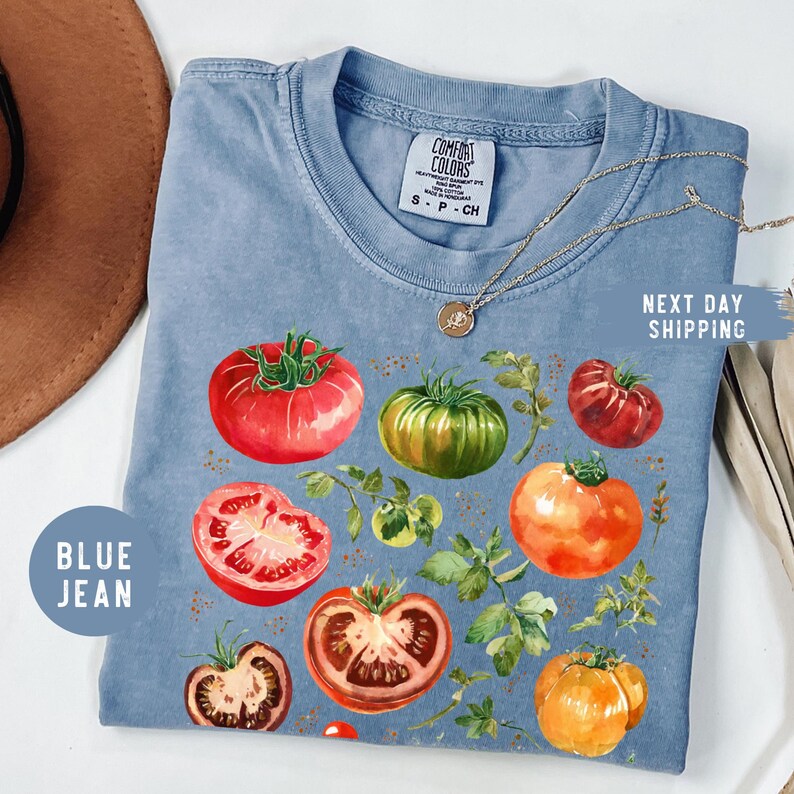 May include: Blue jean Comfort Colors t-shirt featuring a watercolor illustration of various tomatoes in shades of red, green, and orange, with green leaves. The shirt has a crew neck and short sleeves. The text "BLUE JEAN" is on the left side.