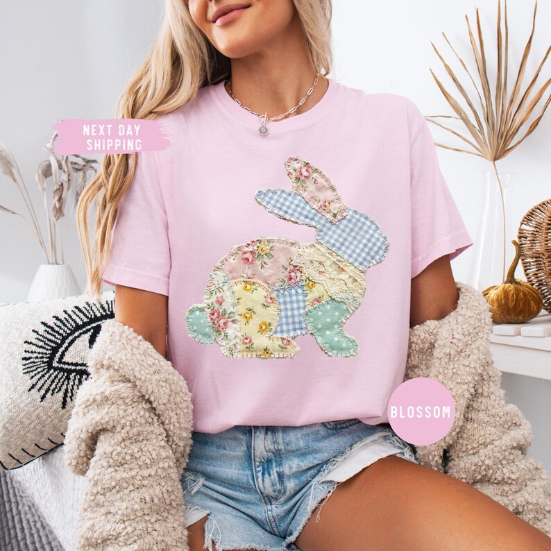 May include: A light pink t-shirt featuring a patchwork bunny design. The bunny is made of floral, gingham, and lace fabrics. The shirt has short sleeves and a crew neck. The text "Blossom" is on the shirt.
