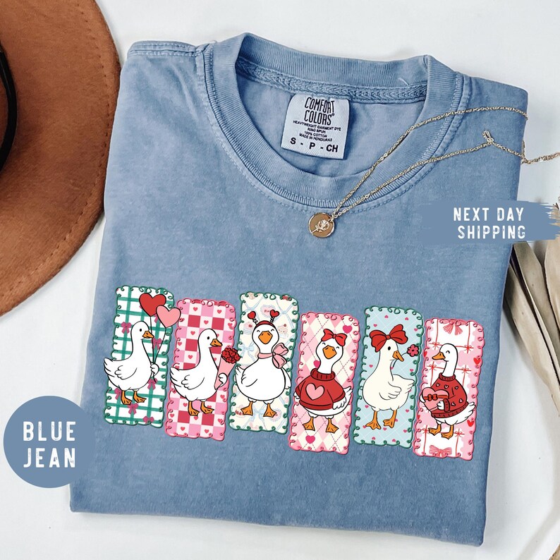 Puede incluir: Camiseta azul vaquero Comfort Colors con dibujos de gansos de dibujos animados con dise&ntilde;os del D&iacute;a de San Valent&iacute;n. Cada ganso tiene un atuendo y un accesorio &uacute;nicos, como un coraz&oacute;n, un lazo o un su&eacute;ter. Un collar dorado est&aacute; drapeado sobre la camisa.