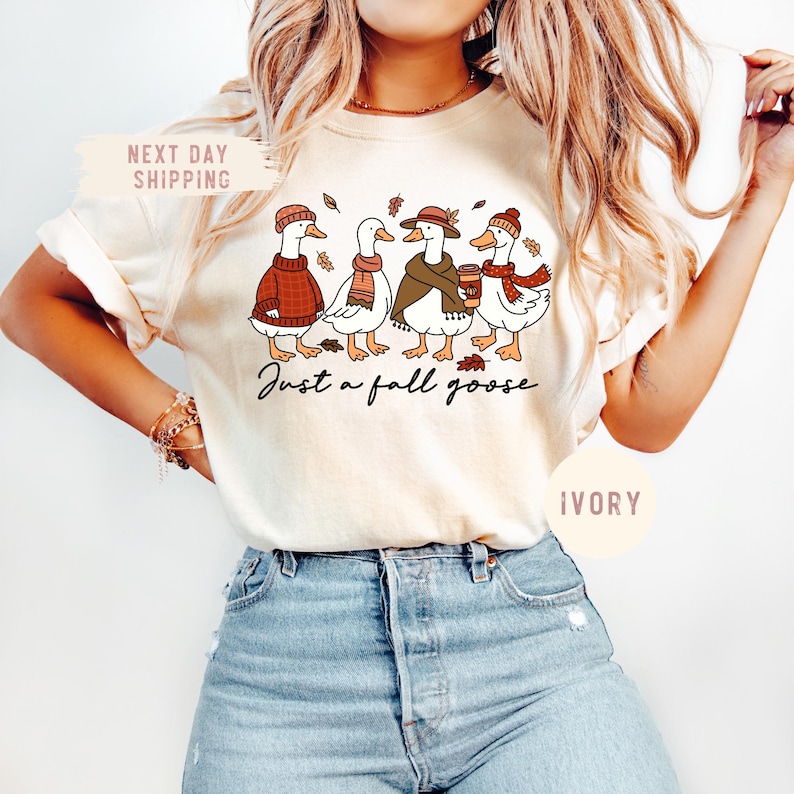 May include: Ivory-colored t-shirt featuring a whimsical illustration of four geese dressed in fall attire. The geese are wearing sweaters, scarves, and hats, with one holding a coffee cup. The text "Just a fall goose" is written below the geese.
