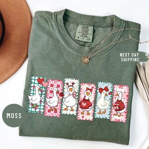 Puede incluir: Camiseta Comfort Colors verde musgo con un gr&aacute;fico de seis gansos de dibujos animados con dise&ntilde;os del D&iacute;a de San Valent&iacute;n. Cada ganso tiene un atuendo y accesorio &uacute;nico, incluyendo corazones y lazos. La camiseta tambi&eacute;n tiene una etiqueta peque&ntilde;a que dice "S P CH".