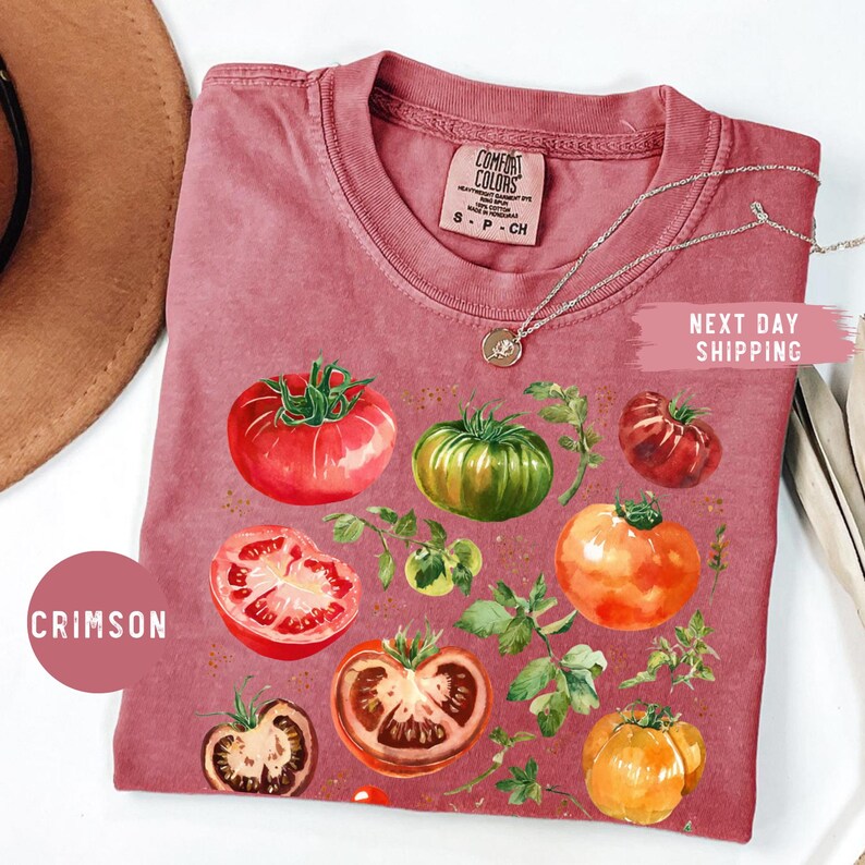 May include: Crimson-colored t-shirt featuring a watercolor illustration of various tomatoes and foliage. The shirt has a crew neck and short sleeves. The text "Comfort Colors" is visible on the tag. A silver necklace is draped across the shirt.