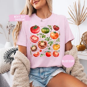 May include: A light pink t-shirt featuring a watercolor design of various tomatoes in shades of red, green, and orange, with green leaves. The shirt has the text "Blossom" on the bottom right and "Next Day Shipping" on the top left.