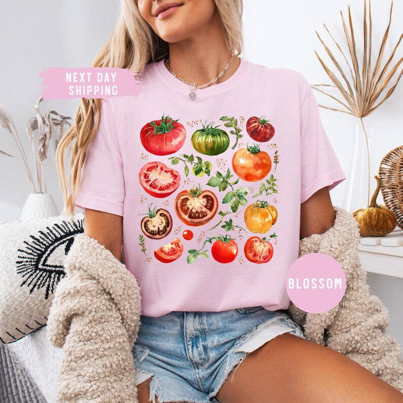 May include: A light pink t-shirt featuring a watercolor design of various tomatoes in shades of red, green, and orange, with green leaves. The shirt has the text "Blossom" on the bottom right and "Next Day Shipping" on the top left.