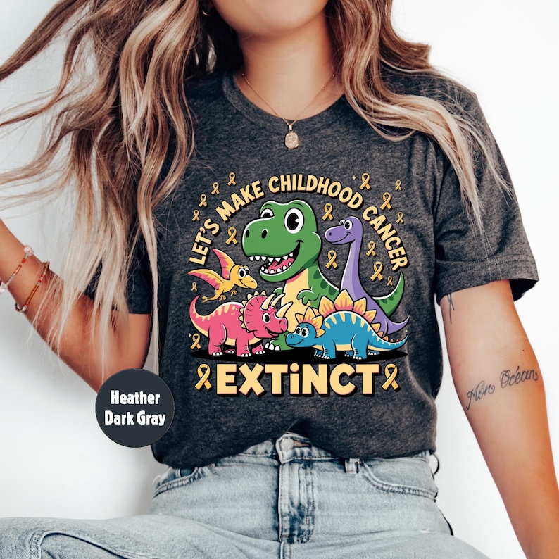 May include: Dark grey heather t-shirt featuring a dinosaur graphic with the text "Let's Make Childhood Cancer Extinct" and yellow ribbons. The shirt has a crew neck design.