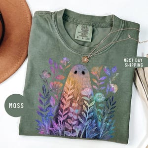May include: Moss green Comfort Colors t-shirt featuring a ghost design surrounded by colorful flowers. The ghost is a gradient of yellow, orange, and pink. The shirt has the text "NEXT DAY SHIPPING" on the right side.
