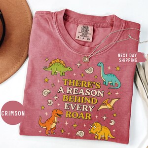 May include: Crimson Comfort Colors t-shirt featuring dinosaur illustrations and the text "THERE'S A REASON BEHIND EVERY ROAR." The shirt has a crew neck and short sleeves. A silver necklace with a pendant is on the shirt.