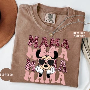 May include: Espresso-colored t-shirt with the word "MAMA" in pink leopard print and a Minnie Mouse graphic wearing heart-shaped sunglasses. The shirt has a crew neck and a Comfort Colors tag.
