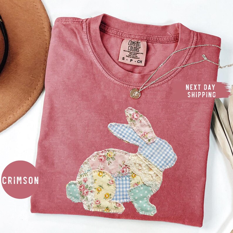 May include: Crimson-colored t-shirt with a patchwork bunny design. The bunny is made of floral, gingham, and lace fabrics. A silver necklace with a pendant is draped on the shirt. The shirt has a Comfort Colors label.