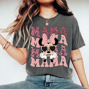 May include: Dark gray t-shirt with the word "MAMA" in pink, leopard print, and a Minnie Mouse graphic. Minnie Mouse is wearing heart-shaped sunglasses and a pink bow. The shirt is a comfortable, casual style.