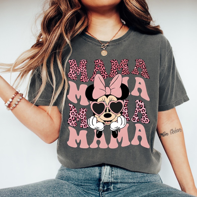 May include: Dark gray t-shirt with the word "MAMA" in pink, leopard print, and a Minnie Mouse graphic. Minnie Mouse is wearing heart-shaped sunglasses and a pink bow. The shirt is a comfortable, casual style.
