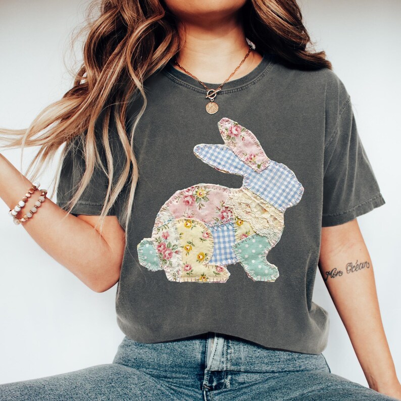 May include: Dark gray t-shirt featuring a patchwork bunny design. The bunny is composed of floral, gingham, and lace fabrics in pastel colors. The shirt has a relaxed fit and short sleeves, perfect for casual wear.