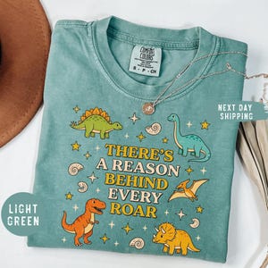 May include: Light green Comfort Colors t-shirt with a dinosaur graphic and the text "THERE'S A REASON BEHIND EVERY ROAR". The shirt features illustrations of various dinosaurs, stars, and shells. A silver necklace is draped across the shirt.