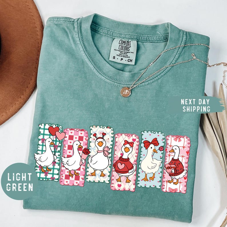 Puede incluir: Camiseta verde claro Comfort Colors con un gr&aacute;fico de seis gansos de dibujos animados con temas de San Valent&iacute;n. Cada ganso est&aacute; en un rect&aacute;ngulo decorativo con corazones y lazos. Un collar dorado est&aacute; sobre la camiseta.