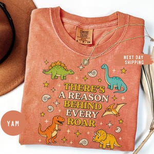 May include: Coral-colored t-shirt with a dinosaur theme. The shirt features various cartoon dinosaurs, stars, and the text "THERE'S A REASON BEHIND EVERY ROAR." A gold necklace and a brown hat are also visible. The shirt has a Comfort Colors tag.