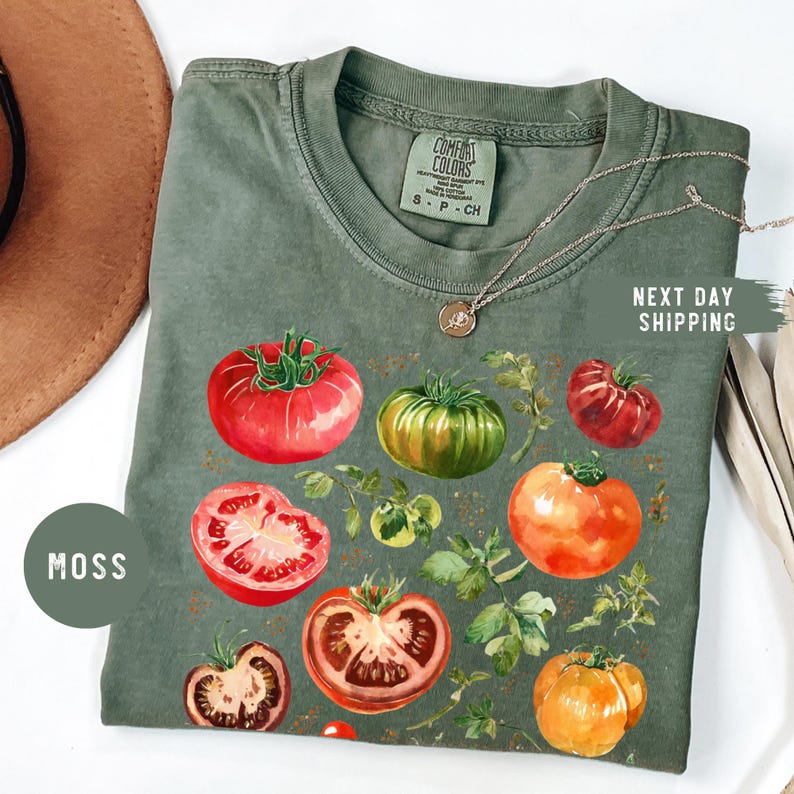 May include: Moss green t-shirt featuring a watercolor design of various tomatoes and foliage. The shirt has a crew neck and short sleeves. The text "NEXT DAY SHIPPING" is visible on the right side of the shirt.