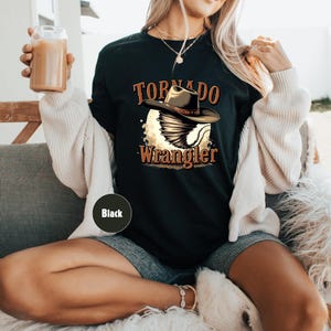 Op de afbeelding: Zwart T-shirt met een afbeelding van een tornado die een cowboyhoed draagt en de tekst "Tornado Wrangler".