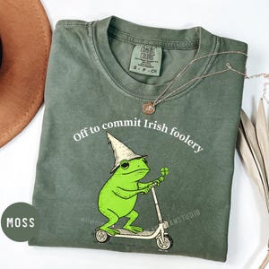 May include: Moss green t-shirt with the phrase "Off to commit Irish foolery" in white. A cartoon frog wearing a hat and holding a four-leaf clover is riding a scooter. The shirt has a Comfort Colors tag.