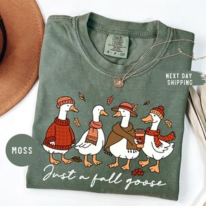 May include: Moss green t-shirt featuring a graphic of four geese dressed in fall attire. The geese are wearing sweaters, scarves, and hats, with one holding a coffee cup. The text "Just a fall goose" is printed below the geese.
