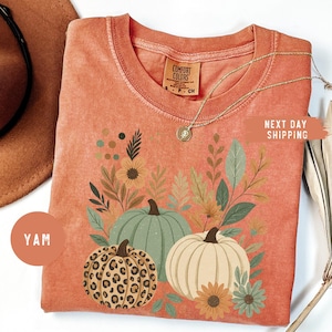 May include: Orange Comfort Colors t-shirt with a fall-themed design. The graphic shows three pumpkins: one with a leopard print, one green, and one cream-coloured, with flowers and leaves. A gold necklace is draped across the shirt.