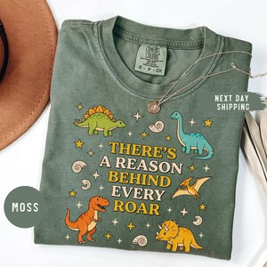 May include: Moss green t-shirt with a dinosaur graphic and the text "THERE'S A REASON BEHIND EVERY ROAR". The shirt features various colorful dinosaurs and stars. The shirt is a Comfort Colors brand.