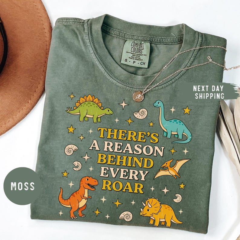 May include: Moss green t-shirt with a dinosaur graphic and the text "THERE'S A REASON BEHIND EVERY ROAR". The shirt features various colorful dinosaurs and stars. The shirt is a Comfort Colors brand.