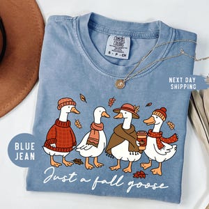 May include: Blue jean-colored t-shirt featuring a whimsical illustration of four geese dressed in fall attire. The geese are wearing hats, scarves, and sweaters, with the text "Just a fall goose" below. A gold necklace and a brown hat are also visible.
