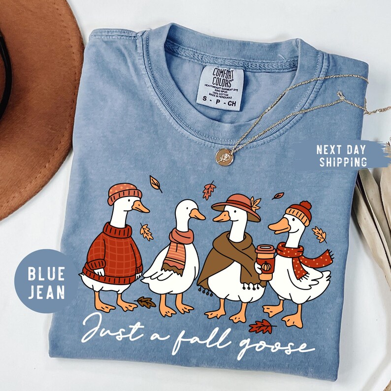 May include: Blue jean-colored t-shirt featuring a whimsical illustration of four geese dressed in fall attire. The geese are wearing hats, scarves, and sweaters, with the text "Just a fall goose" below. A gold necklace and a brown hat are also visible.