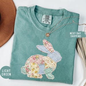 May include: Light green Comfort Colors t-shirt with a patchwork bunny design. The bunny is made of floral, gingham, and polka dot fabrics. A silver necklace with a pendant is draped on the shirt. The shirt has the text "Light Green".