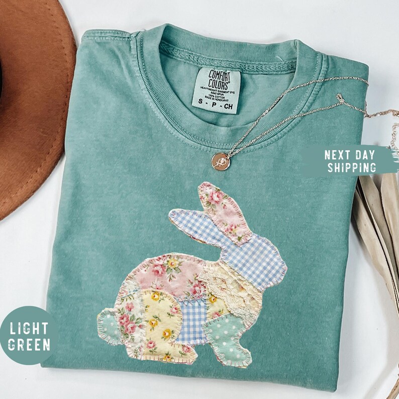 May include: Light green Comfort Colors t-shirt with a patchwork bunny design. The bunny is made of floral, gingham, and polka dot fabrics. A silver necklace with a pendant is draped on the shirt. The shirt has the text "Light Green".