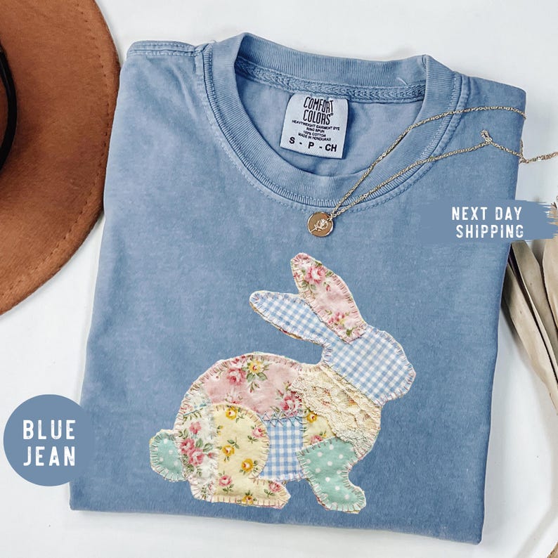 May include: A blue jean Comfort Colors t-shirt featuring a patchwork bunny design. The bunny is made of floral, gingham, and polka dot fabrics. A gold necklace with a pendant is draped on the shirt. The shirt has the text "Next Day Shipping".